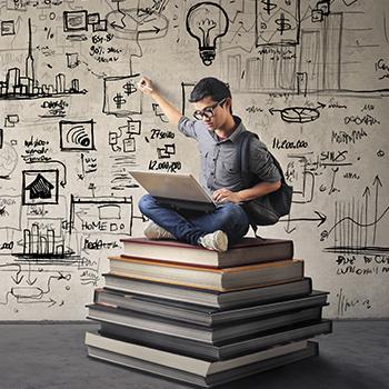Man sitting on large books and holding laptop while pointing to background with lightbulb, graphs and mindmaps.