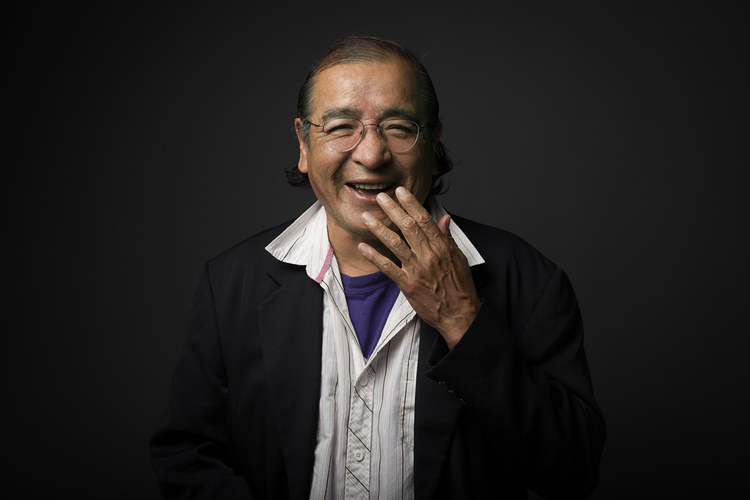 smiling Indigenous man wearing casual jacket and shirt, with hand held to mouth in laughter