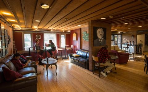 The Trent at Traill College. A warmly lit, pub-like space with tables and chairs, leather couches, and art on the walls. There is a light wooden floor and exposed brick walls. A few students are sitting at the tables.