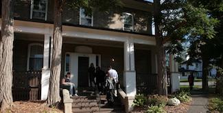 Crawford House, an old, beige brick home with a large covered porch running along its entire front. A few students are sitting on the porch and walking up the front steps.