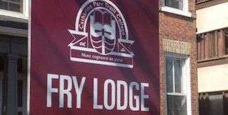 The sign outside Fry Lodge. It is maroon with white lettering and a white Traill College logo. Behind the sign is an old, red-brick home.