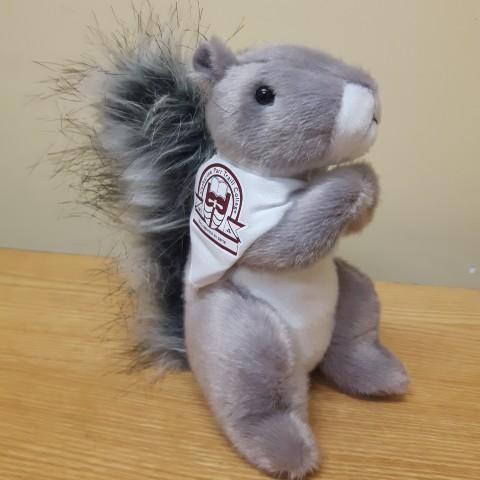 A grey plush squirrel wearing a bandana around its neck. The bandana is white with a maroon Traill College logo.