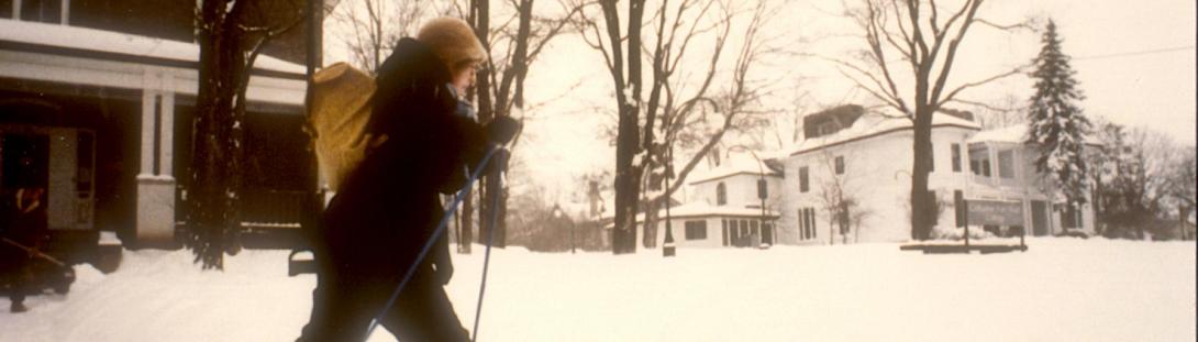 A vintage photograph of a person cross-country skiing past Scott House at Traill College. Snow is covering the ground and the bare tree branches.