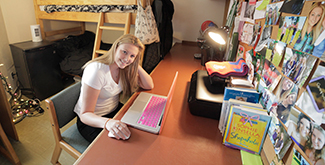 Student sitting at her desk in her dorm room typing on her computer, smiling at the camera