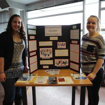 Two girls standing in front of a poster with the title "Occupational Gender Stereotypes"