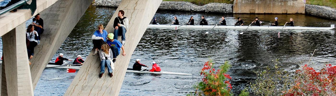 Rowers racing underneath a bridge while students attempt to watch from the bridge supports in a dangerous manner