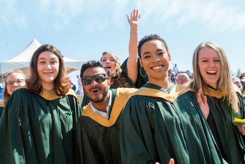 A group of joyful graduates in green robes and hoods celebrates at an outdoor graduation ceremony under a clear blue sky.