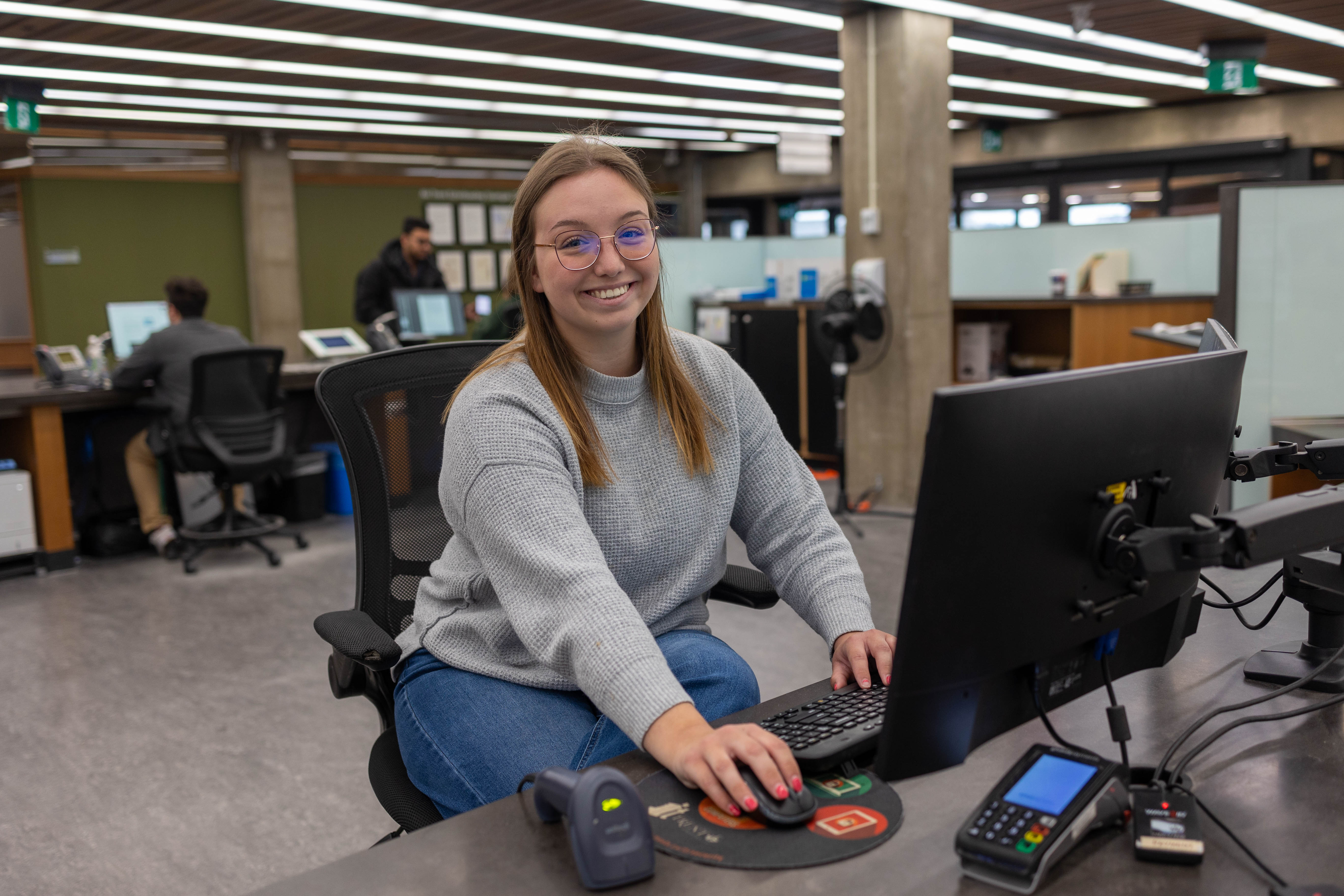 A smiling young woman wearing glasses and a grey sweater sits at a library information desk, working on a computer.