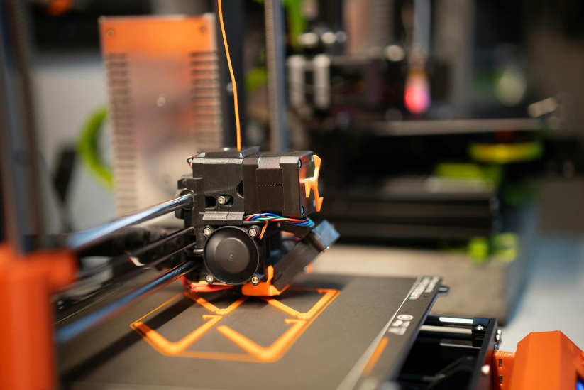 A 3D printer in action, printing an orange object on a heated bed inside a modern workshop.