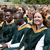 Students sitting in chairs outside on the podium wearing green gowns, smiling at the camera