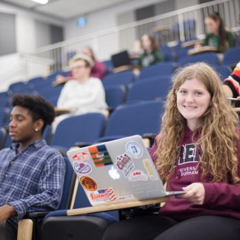 Student with laptop, sitting in a lecture hall smiling, surrounded by other students