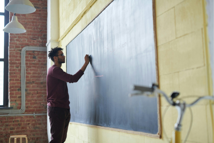A young man in a maroon sweater writes on a large blackboard in a bright, brick-walled classroom, with a bicycle parked to the side.