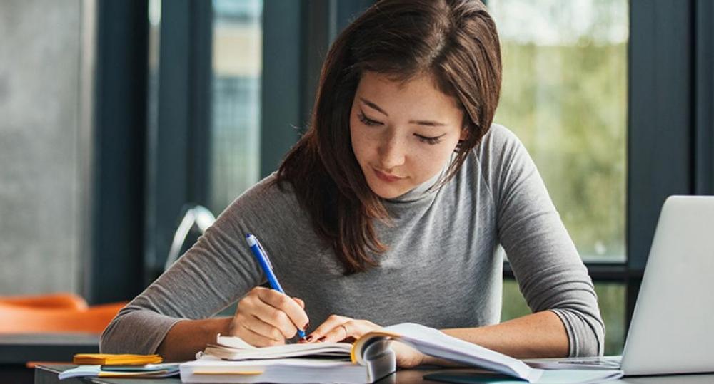 student writing on paper in front of laptop