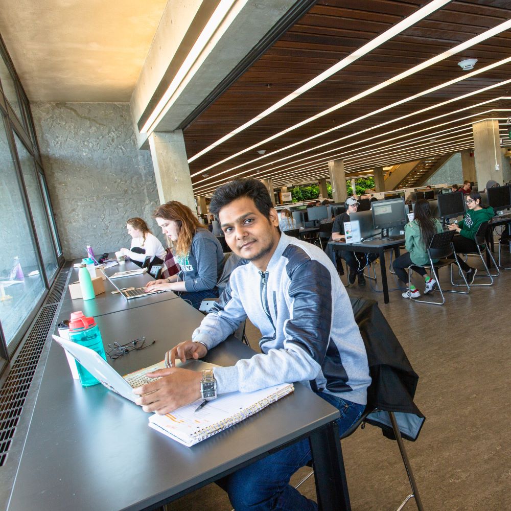 A student sitting at a desk with an open laptop and notebook in Bata Library, surrounded by other students studying alone or in groups 
