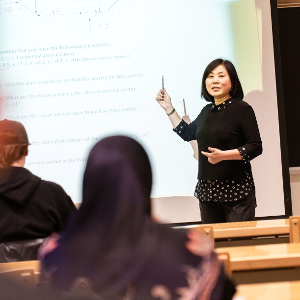 A professor facing a classroom with one hand pointing at a display screen. 