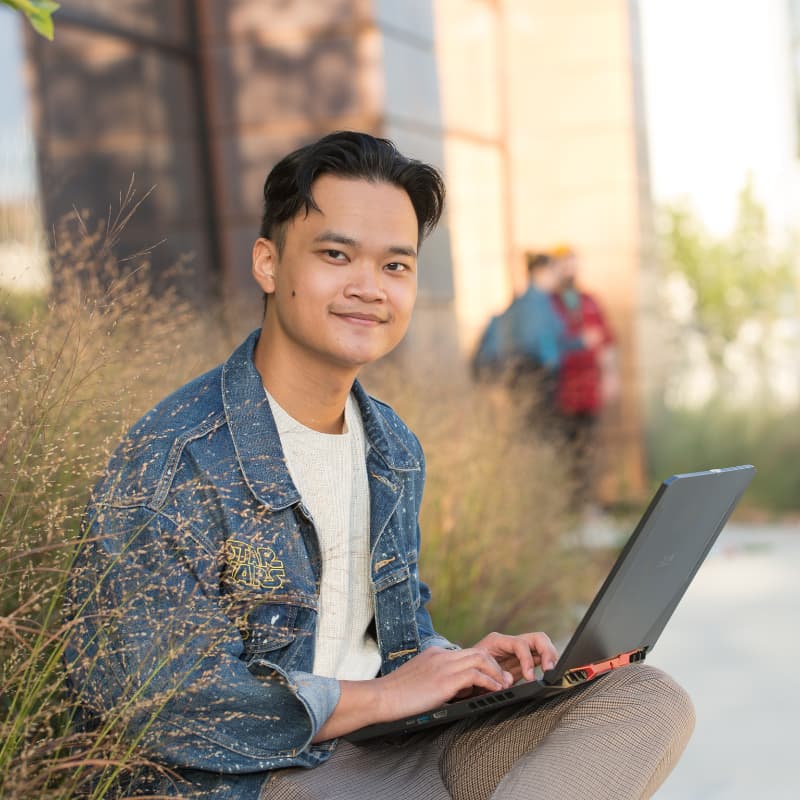 Photo of student working on laptop outside smiling at the camera