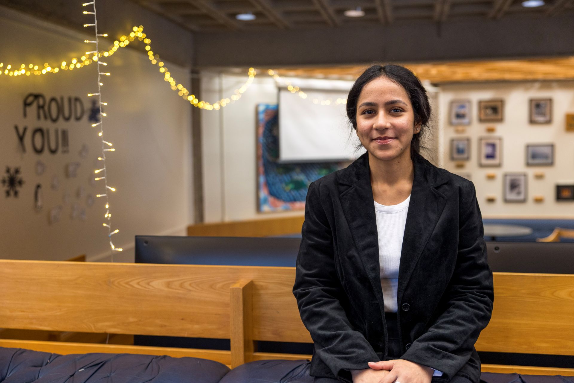A student in a black blazer seated in a student space, facing the camera and smiling.