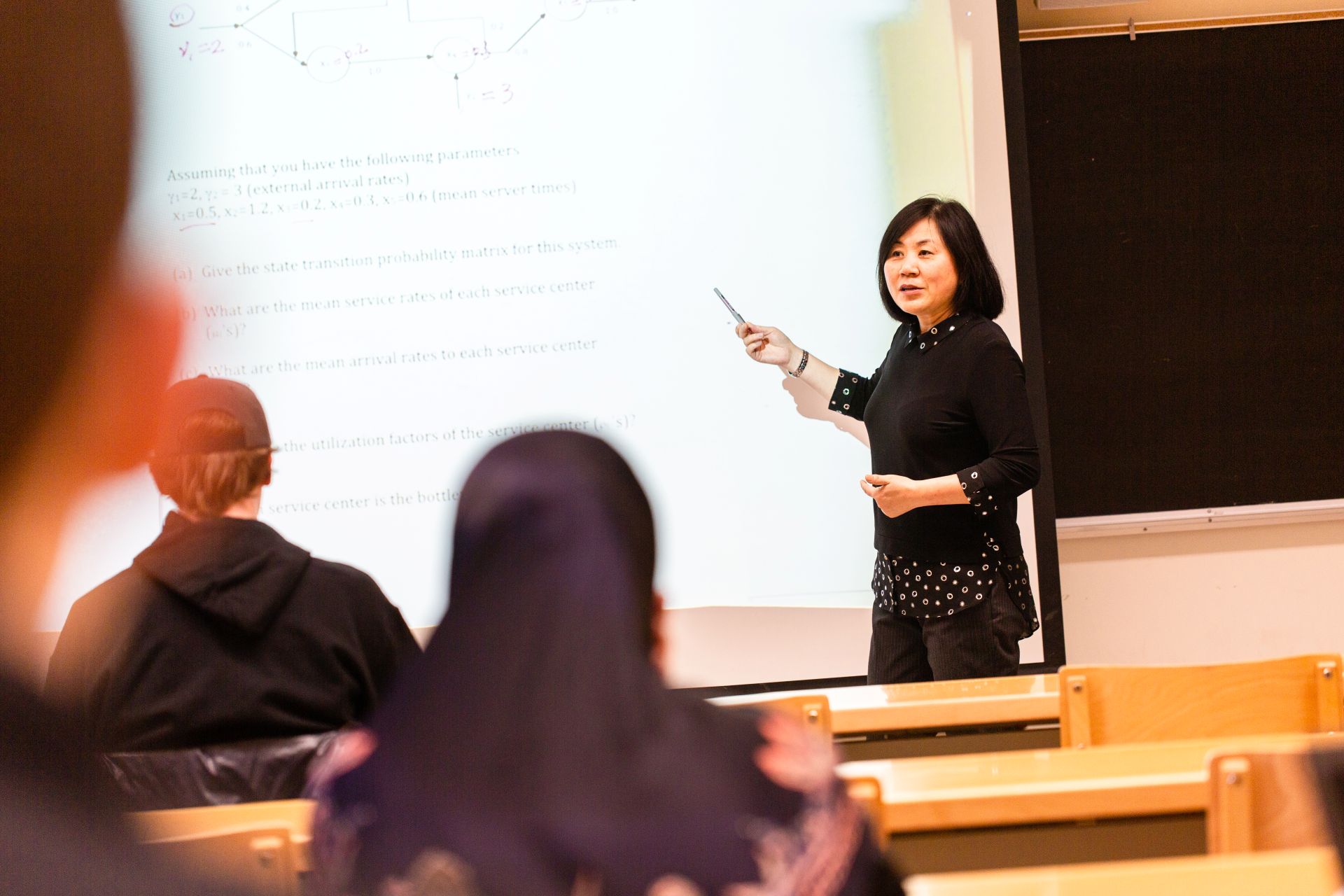 A professor facing a classroom, pointing to a  screen