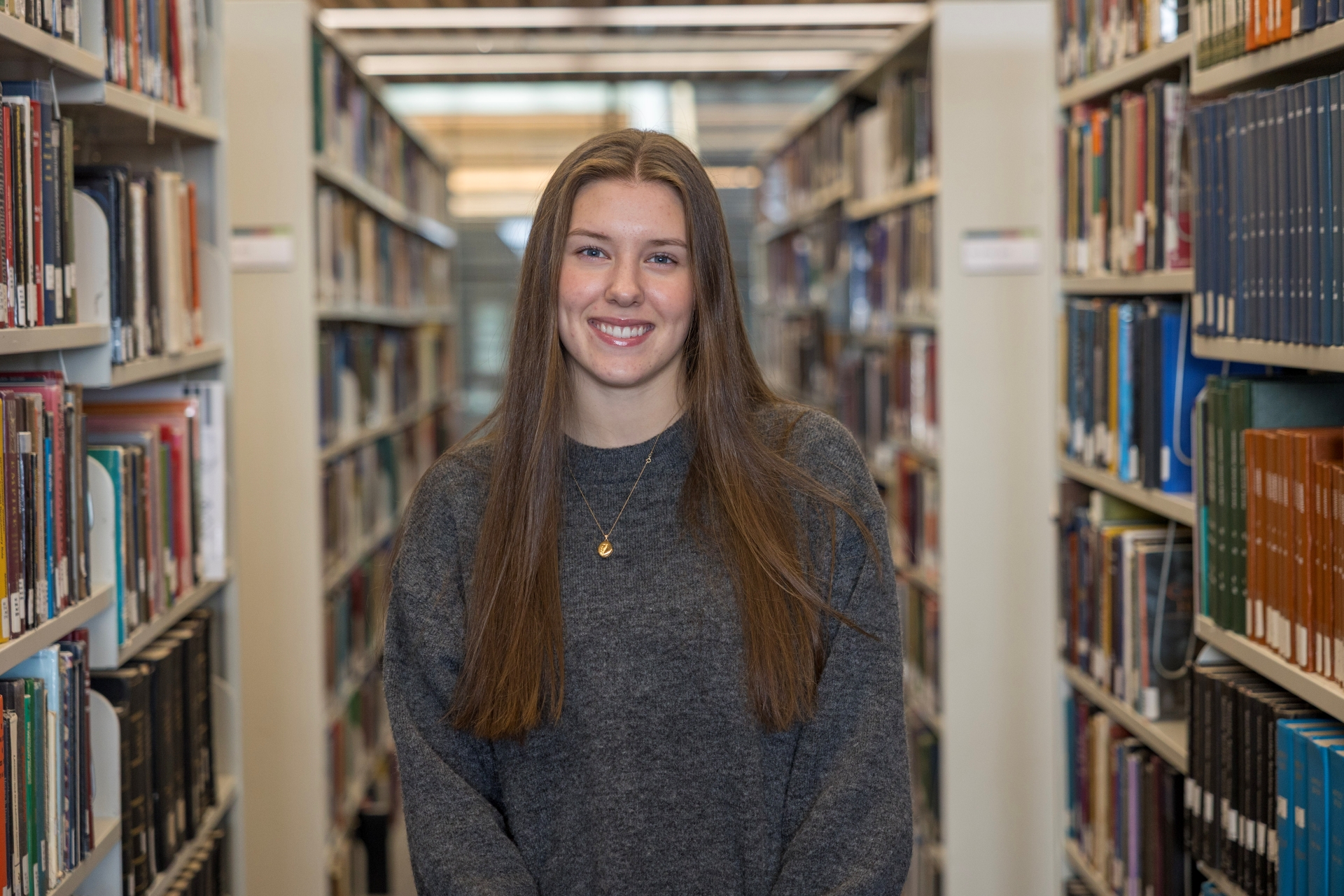 A smiling student facing the viewer, standing in between stacks of books in the library.