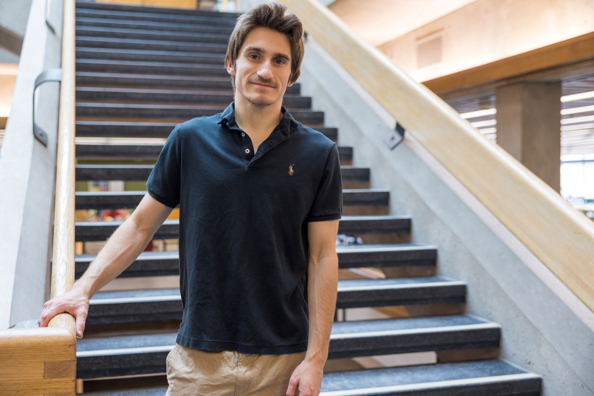 Student in a dark polo shirt facing the camera, smiling, standing at the base of the stairs at Bata Library.