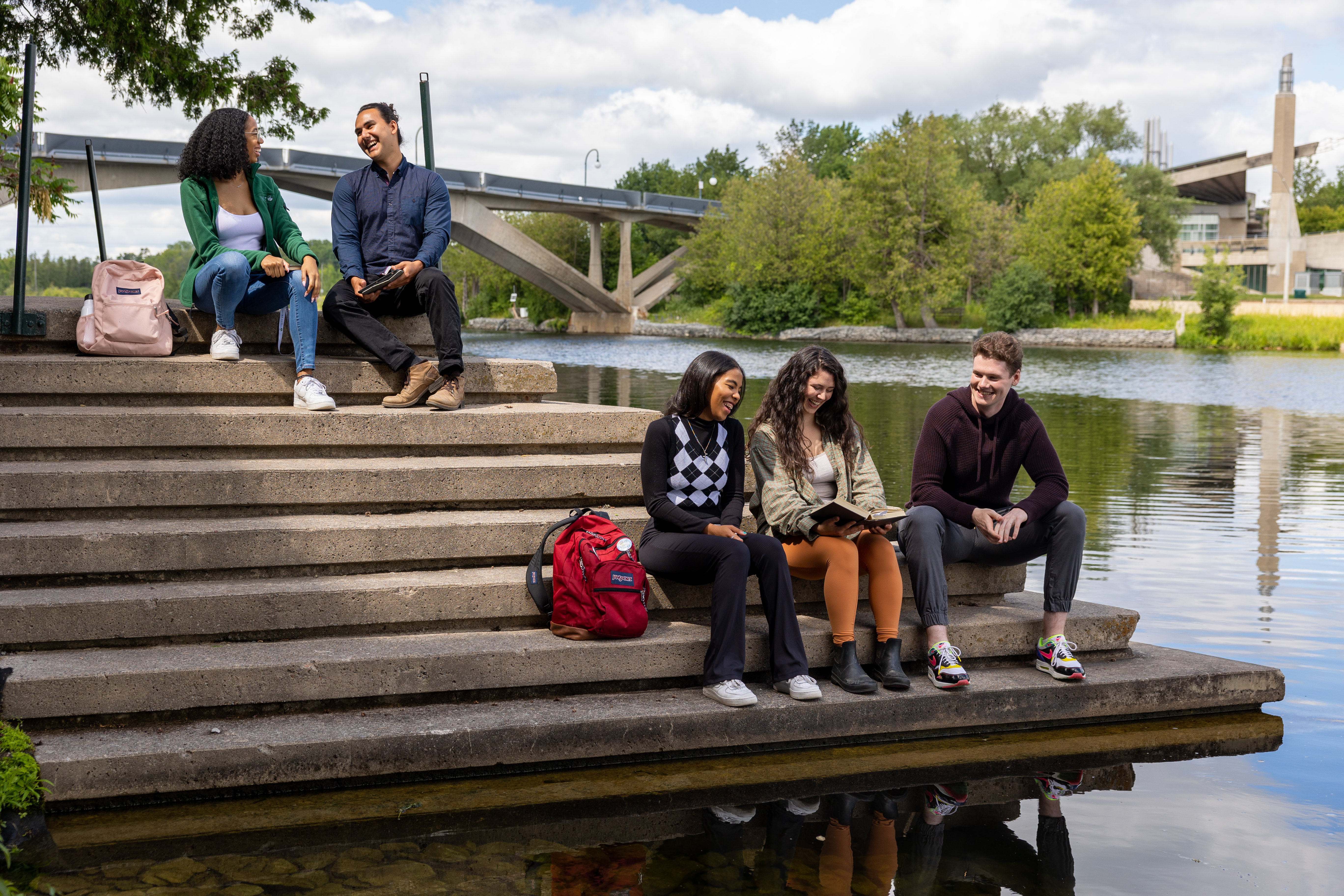 Students sitting on the steps by the Otonabee River, chatting and looking at a book.