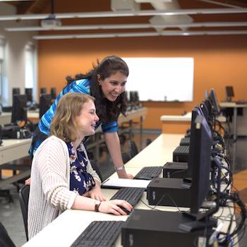 A professor leaning down next to a student working on a computer in a lab
