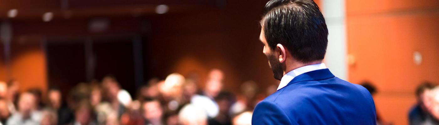 blue suit talks to a crowd of people