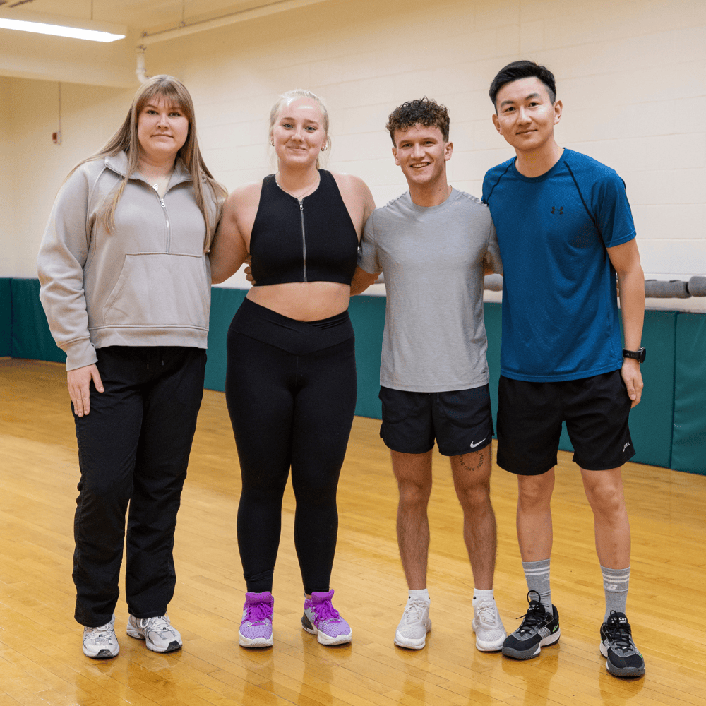 Four people stand in a line in a fitness studio. they are all smiling.