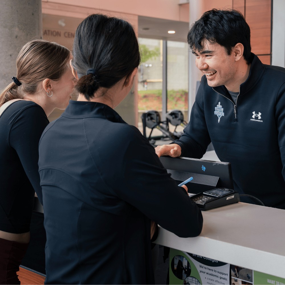 Male employee in a Trent Athletics Centre uniform shirt points to paperwork at the front desk, helping two prospective members. Prospective members have their backs to the camera.