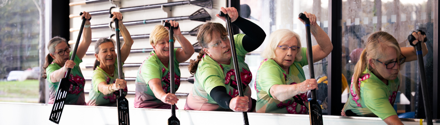Six members of Survivors Abreast in their green and pink jerseys sit in the row tank actively paddling.