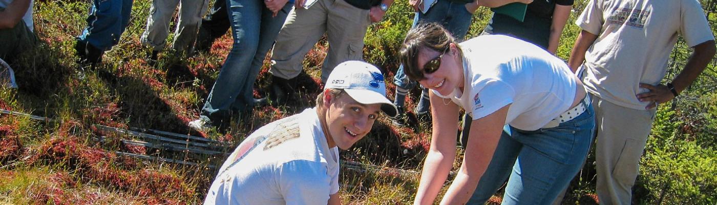 2 students kneeling on the ground holding onto a metal rod pushed into the earth, infront of a row of students watching, in a sunnt woodland area in the summer sun