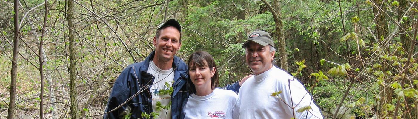 3 faculty members stading beside each other, smiling at the camera in a woodland setting in the summer shade