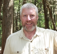 David Beresford standing in a forest in the dappled sun, wearing an off-white shirt and smiling at the camera.