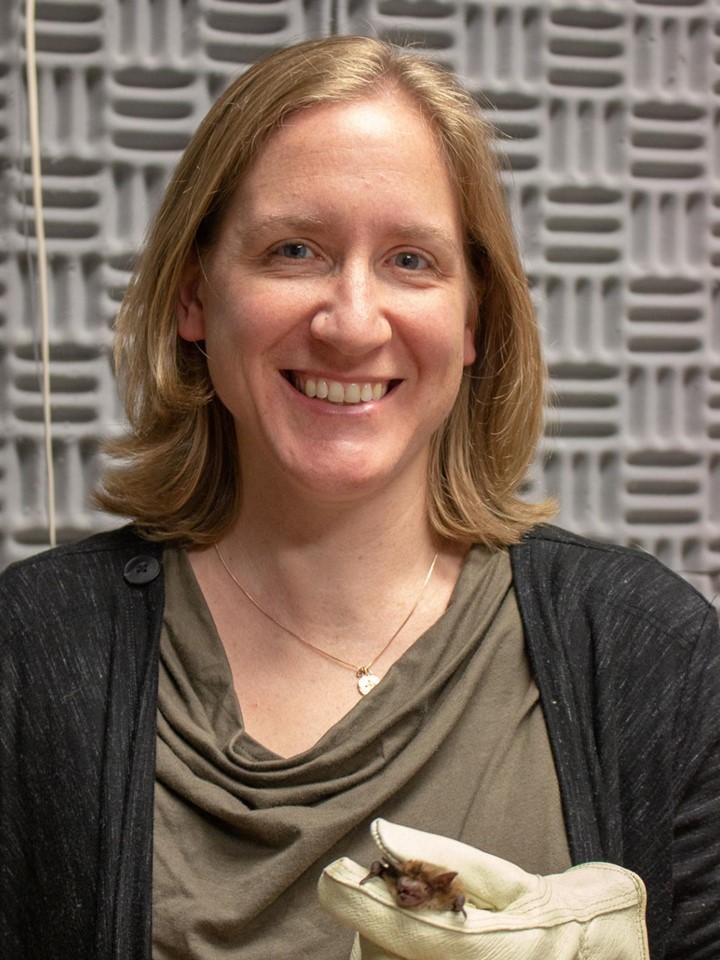 Christina Davy, smiling at the camera while holding a bat in her gloved hand, in front of a sound-stopping wall
