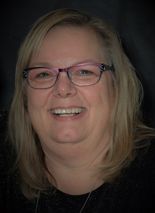 Debbie Lietz wearing purple glasses and smiling at the camera on a dark background