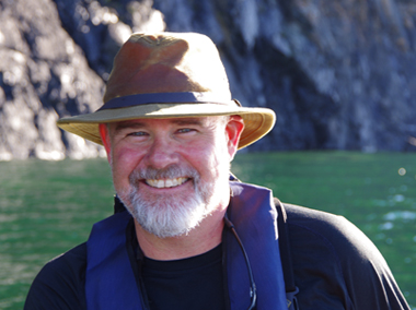 Nick Jones, wearing a brimmed hat in the sun, on the water, smiling at the camera in from of a rock wall
