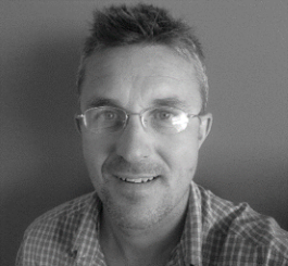 Kirk Hillsley in black and white, wearing glasses and a checkered shirt smiling at the camera.