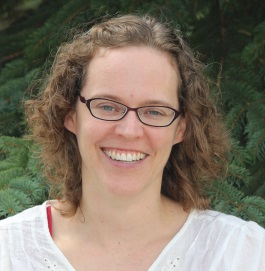 Kelly Boadway in front of a spruce tree wearing glasses and a white shirt, smiling at the camera.