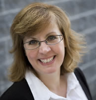 Leslie Kerr standing in front of a dark grey brick wall, wearing a white shirt and black suit jacket, smiling at the camera.