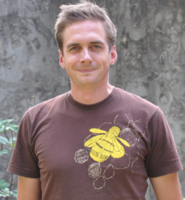 Marcel Dorken standing in front of the cement wall of Champlain College, wearing a brown shirt with a yellow bee on it, smiling at the camera.
