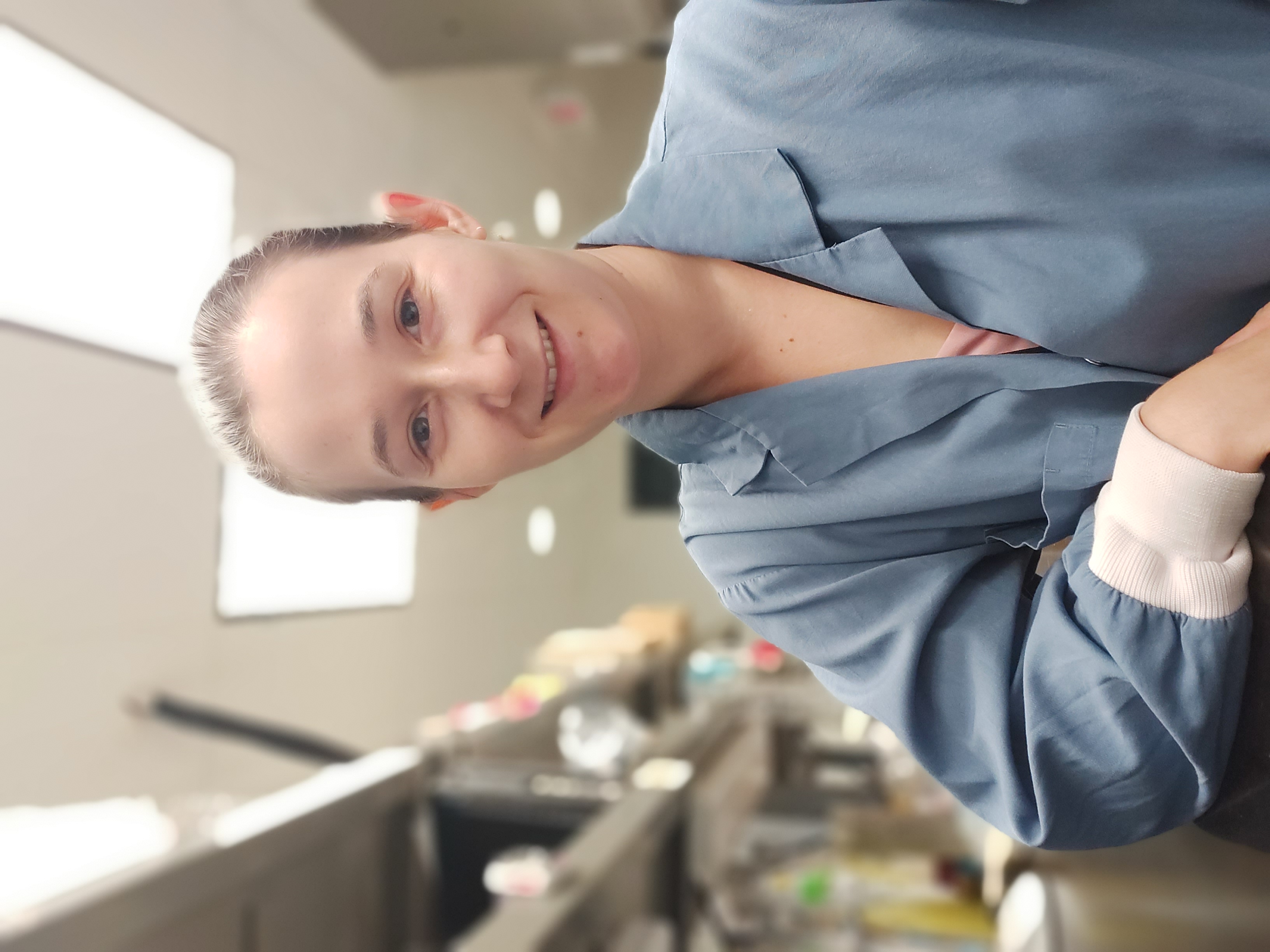 Stephanie Tobin sitting with her right arm on a lab bench, wearing a blue lab coat, and smiling at the camera. 
