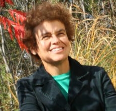 Erica Nol standing in a shrubby meadow on a sunny day with red sumac and long grasses, wearing a black suit jacket and a green shirt, looking up and smiling.