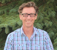 Gary Burness in front of a spruce tree, wearing a blue and purple checkered shirt and glasses, smiling at the camera.