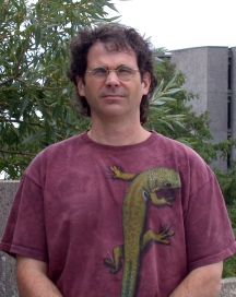 Dennis Murray standing in front of trees and a building wearing glasses and a burgundy shirt with an iguana on it, squinting at the camera.
