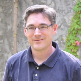 Paul Frost standing in front of the cement wall of Champlain College and a green leafy vine growing on it, wearing glasses and a navy blue shirt, smiling at someone behind and to the left of the camera.