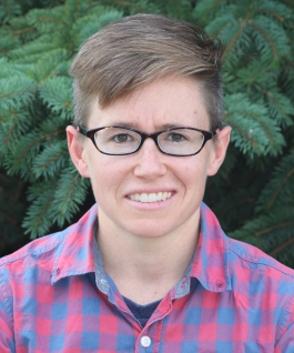 Sara Pieper in front of a spruce tree wearing glasses and a dark pink and blue checkered shirt, smiling at the camera.