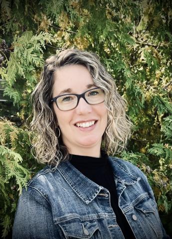 Photo of a woman in glasses smiling in front of green shrubs