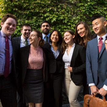 A group of students standing side by side agaist a brick wall covered in green ivy in the summer shade, wearing business suits
