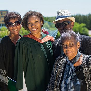 A female student in her graduation gown and copper hood standing on the Faryon bridge withe 5 family members, smiling at the camera in the summer sun
