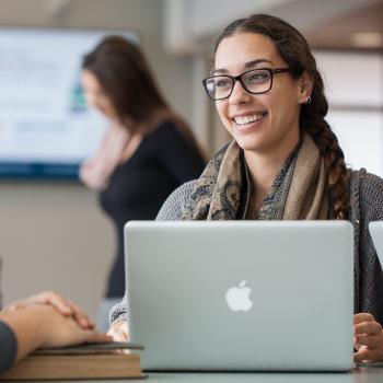 A female student sitting in the atrium talking to another student in front of a laptop